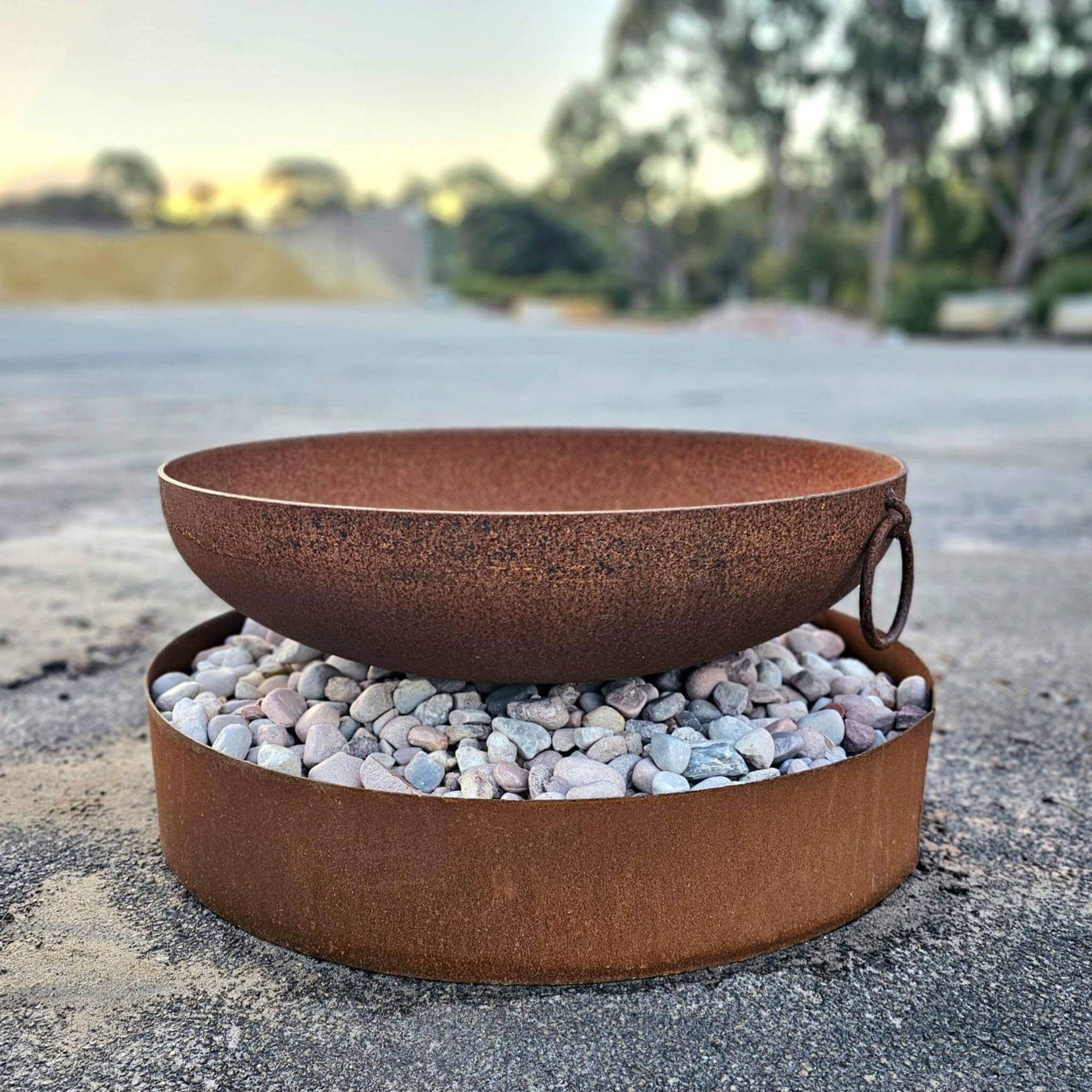 A rusty fire bowl with handles sitting atop a corten steel ring filled with river stone at Designer Dirt in Albany